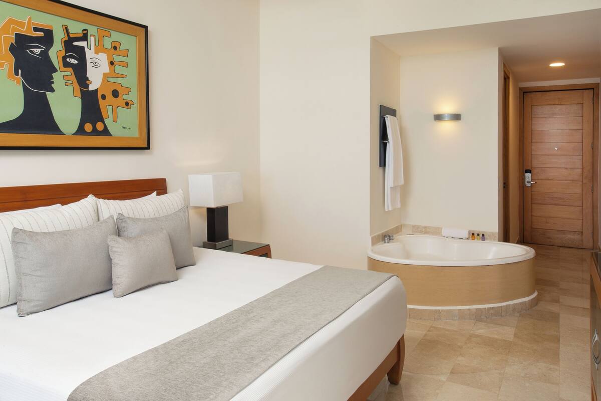 Une chambre d’hôtel avec un grand lit, une table de chevet avec une lampe, un tableau au mur et une salle de bain séparée avec une baignoire.