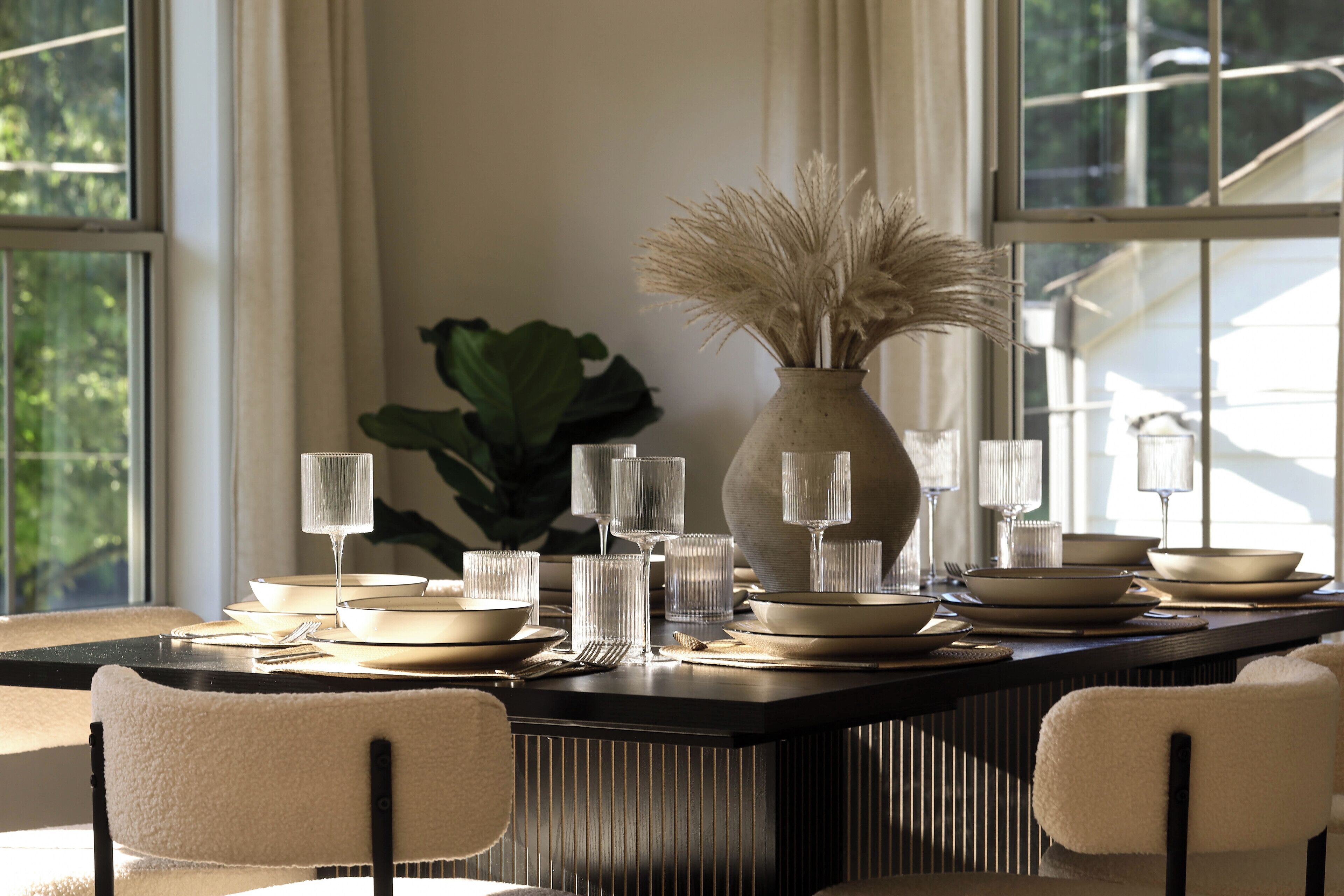 Un coin repas avec une table rectangulaire dressée pour quatre personnes, agrémenté d’un vase contenant des herbes de la pampa séchées, de verres transparents et d’assiettes.