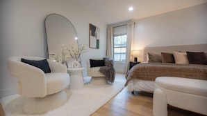 Un dormitorio moderno con una cama grande, dos sillones blancos, una mesa redonda, una lámpara de pie y un ventanal con cortinas.