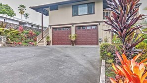 Une maison à deux étages avec un garage, entourée d’un jardin aménagé avec différentes plantes et un palmier.