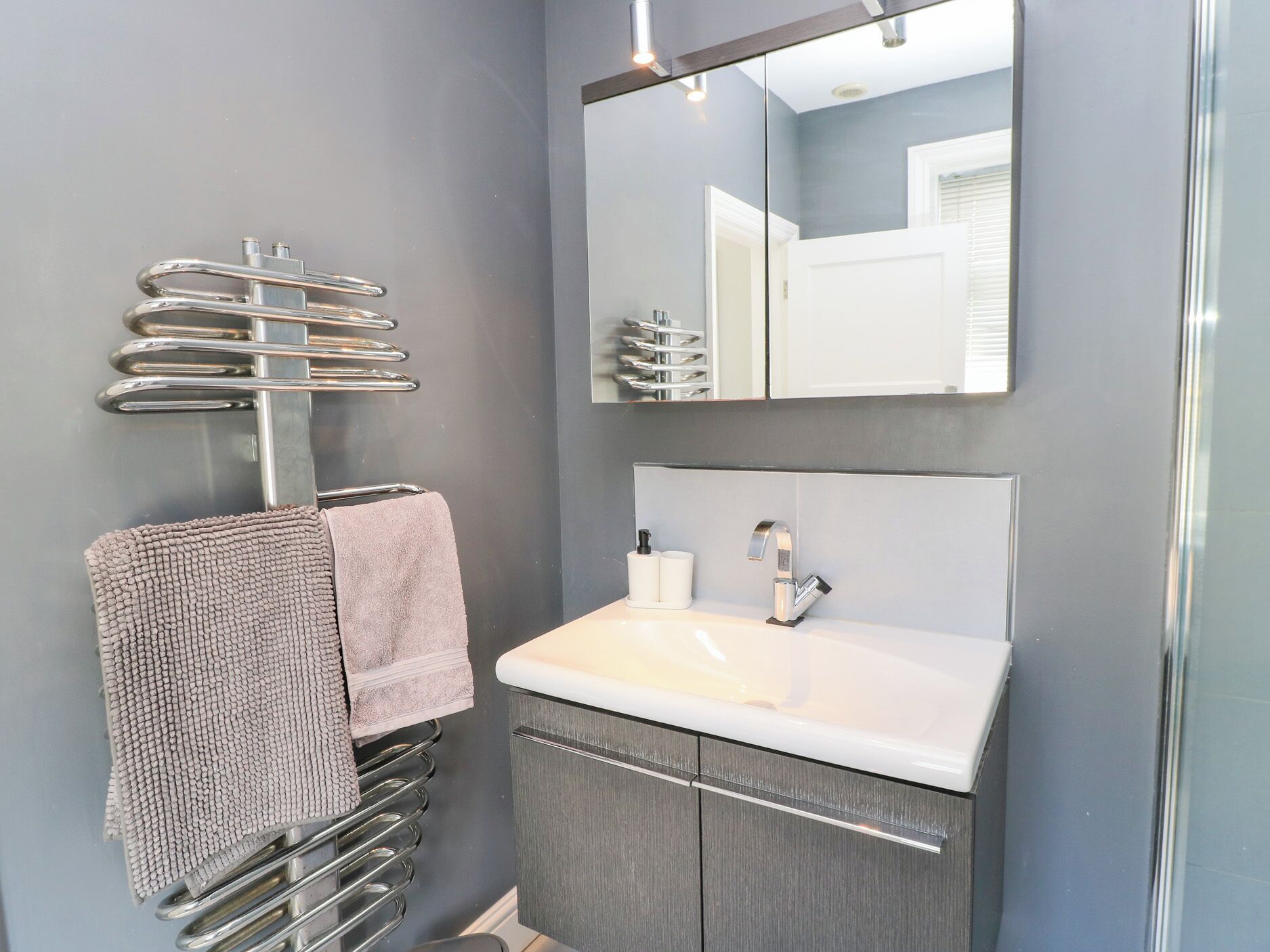 Une salle de bain moderne avec un mur gris, un lavabo blanc, un grand miroir et un porte-serviettes sur lequel est posée une serviette rose.