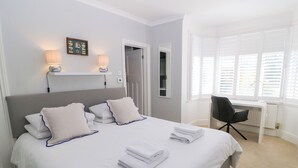 Ein ordentlich bezogenes Bett mit weißen Leinen und blauer Biese, ein Schreibtischstuhl und ein Fenster mit Jalousien.