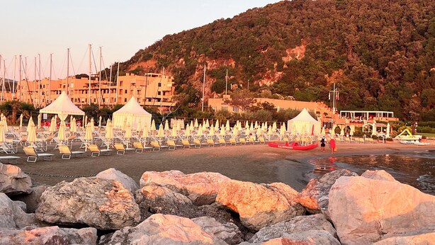 Ein Strand mit Reihen von Liegestühlen und Sonnenschirmen, einem felsigen Vordergrund und Gebäuden im Hintergrund.