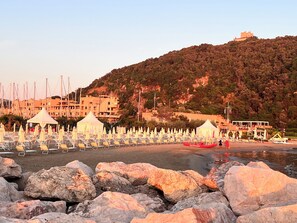 A beach with rows of sun loungers and umbrellas, a rocky foreground, and buildings in the background.