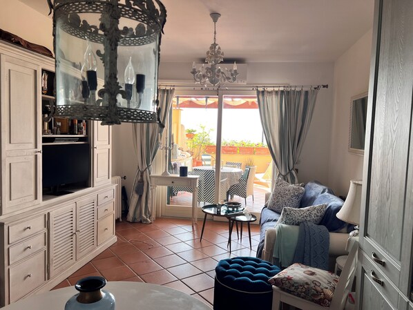 A well-lit living area with a sofa, ottoman, and a dining table. A large window offers a view of the outdoors.