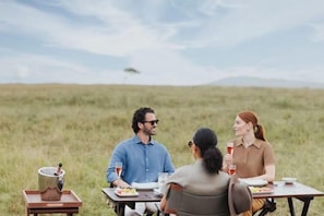 Tres personas cenando en una mesa al aire libre en un campo cubierto de hierba, con cielo despejado.