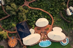 Vista aérea de un complejo turístico con un área de piscina central, múltiples zonas de estar con sombrillas y una terraza de madera circular.