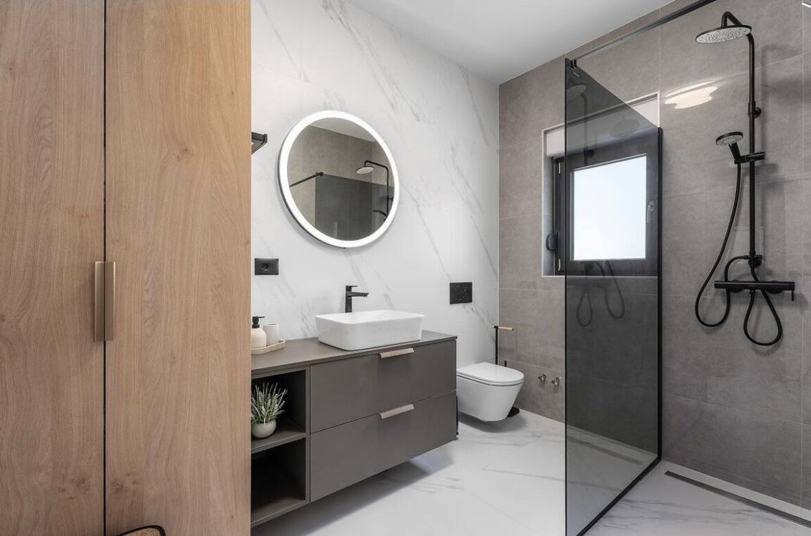 A modern bathroom with a wooden vanity, a round mirror, a white sink, a toilet, and a glass-enclosed shower area.