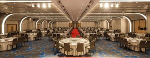 A large banquet hall with multiple round tables and chairs, a red carpet, and a modern ceiling design.