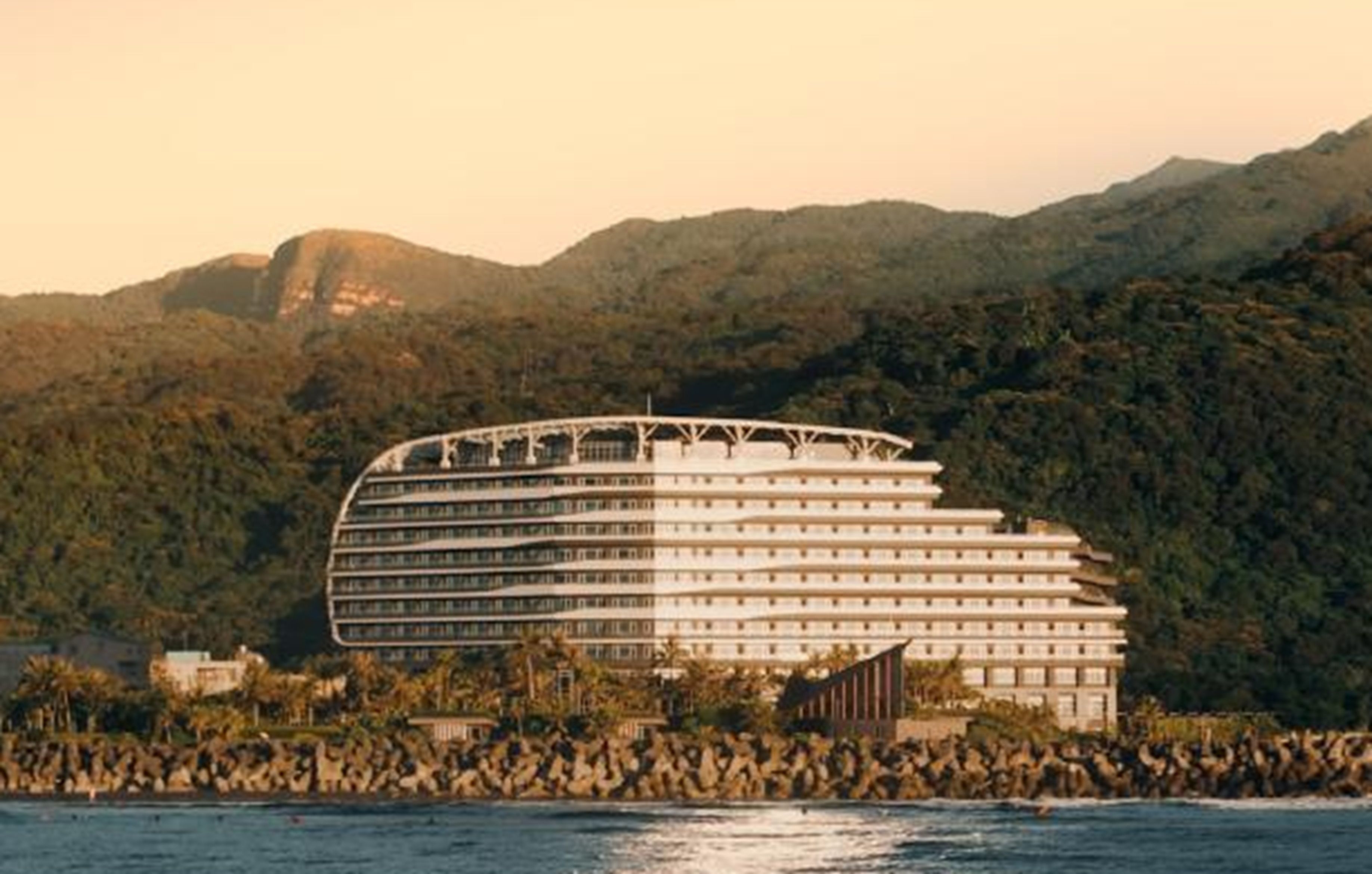 A modern hotel with a curved facade, located near a body of water with mountains in the background.