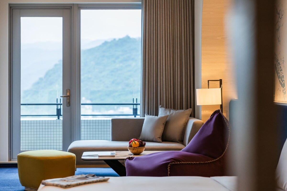 Una camera d'albergo con un divano, un piccolo tavolo con una ciotola di frutta, una lampada e vista sulle montagne tramite una porta scorrevole.