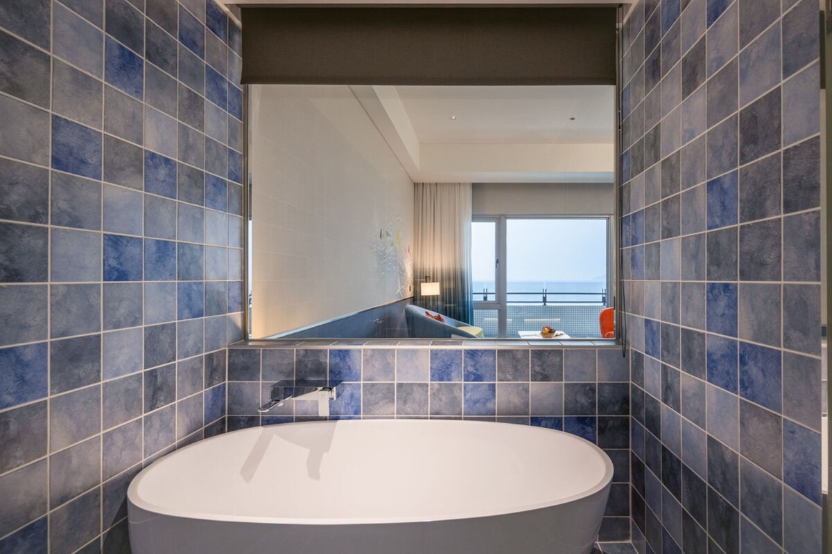 Un bagno moderno con una vasca da bagno ampia, pareti rivestite di piastrelle blu e vista su un balcone attraverso un grande specchio.