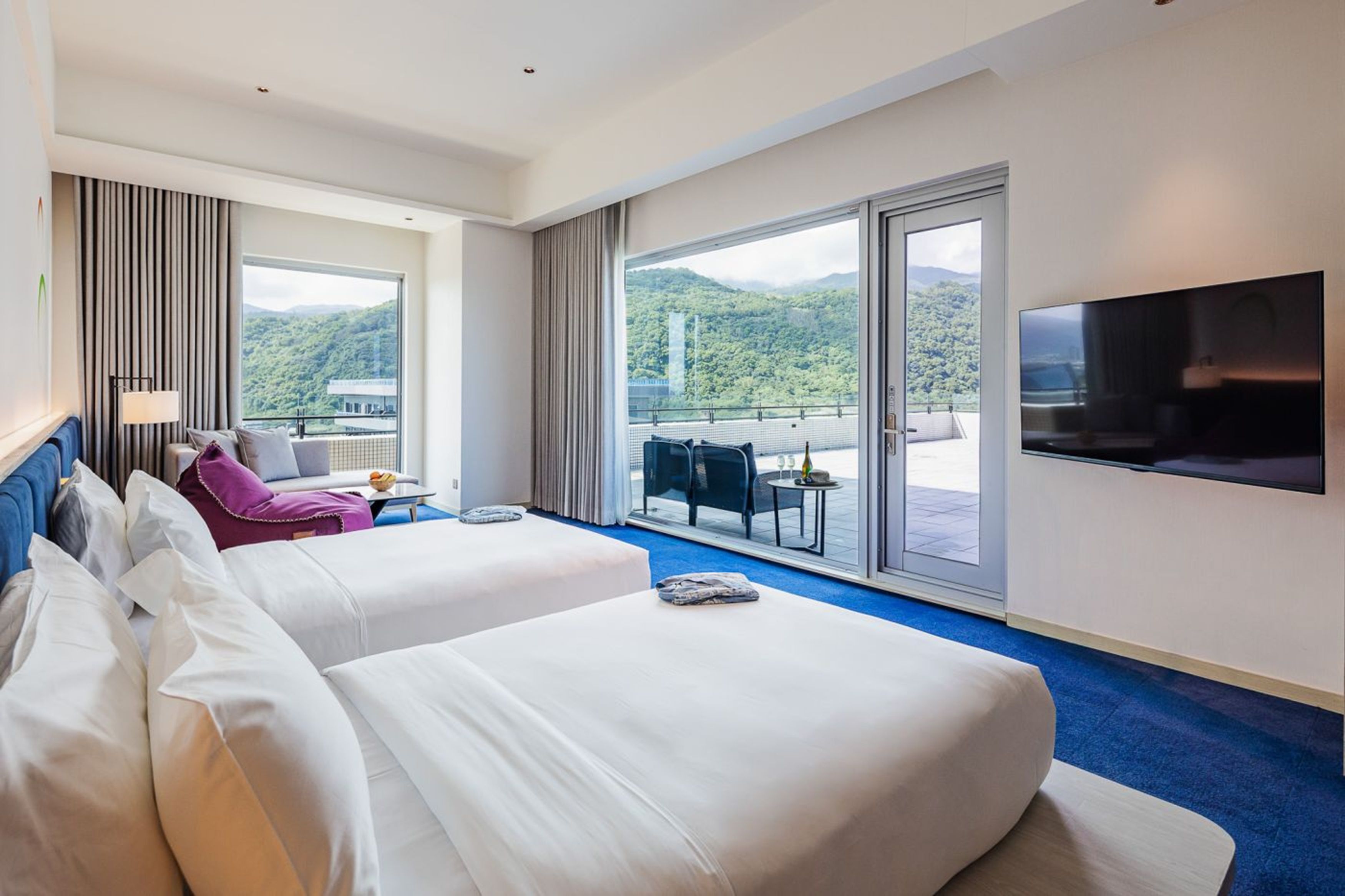 A hotel room with two beds, a TV, a balcony with a table and chairs, and a view of a mountainous landscape.