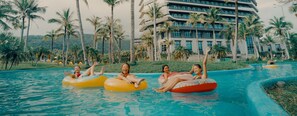Une piscine où des personnes flottent sur des bouées gonflables, entourée de palmiers et d’un grand bâtiment.