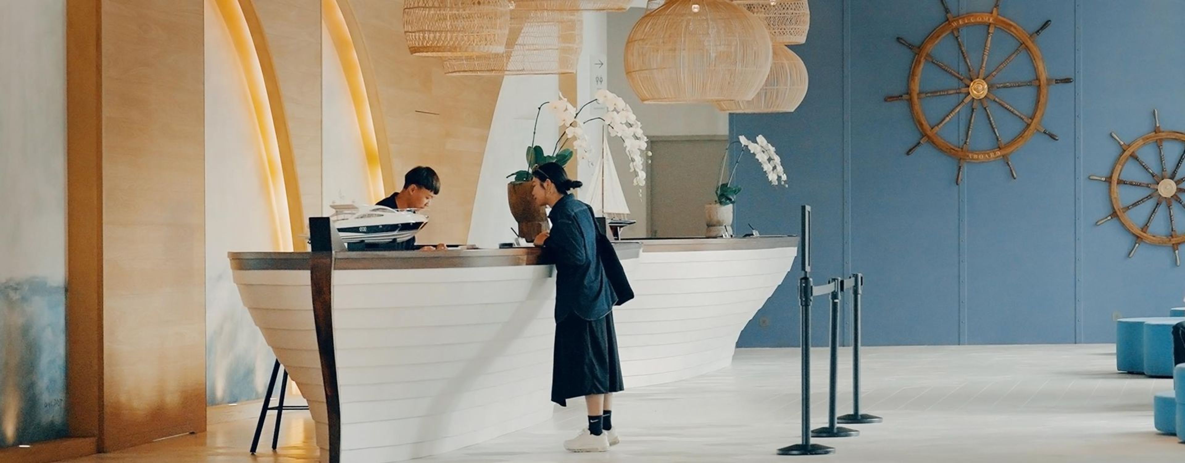 A reception desk with a person standing behind it, a ship wheel on the wall, and a potted plant.