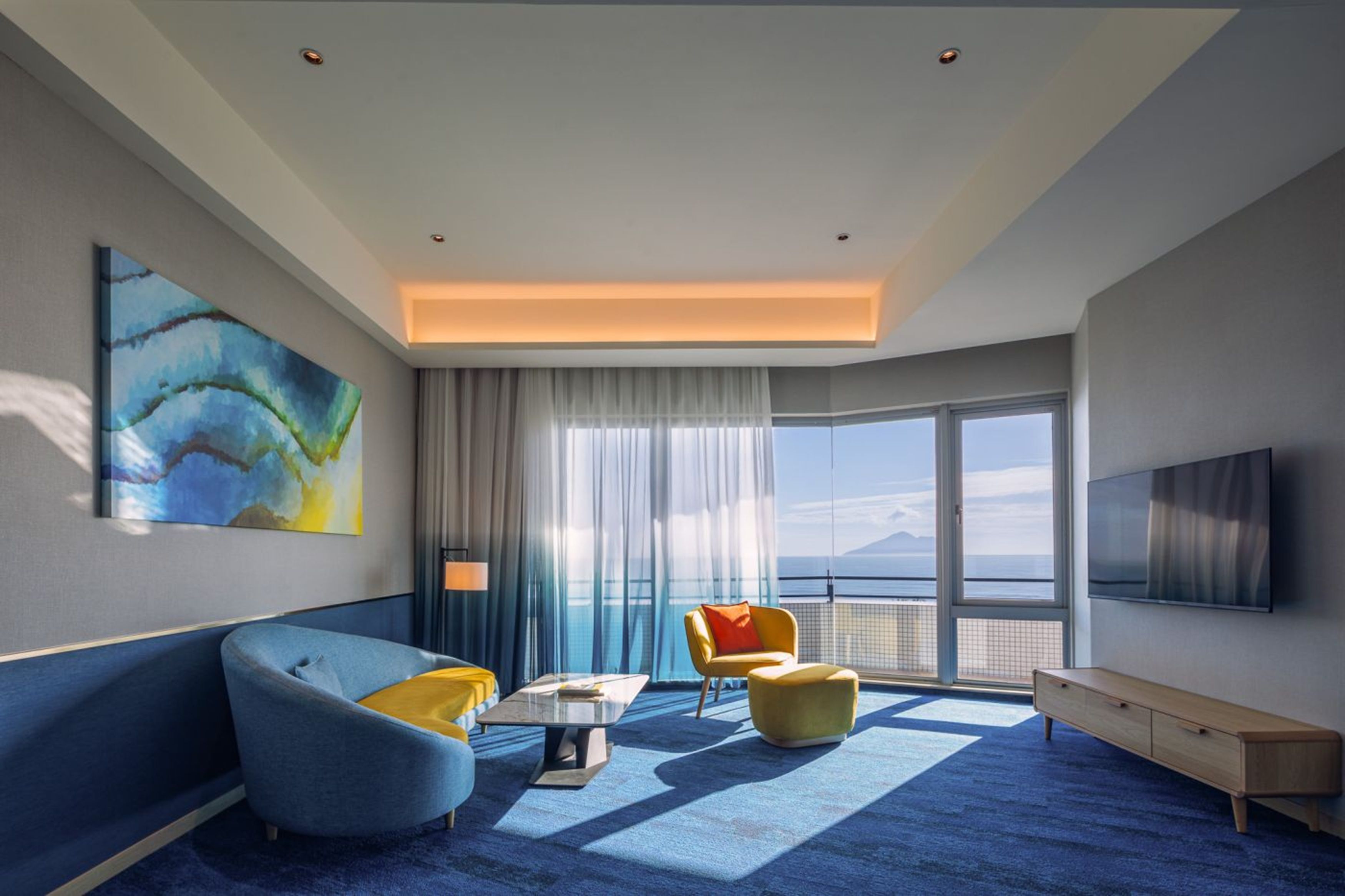 A modern hotel room with a large window, a sofa, a chair, a coffee table, and a TV.