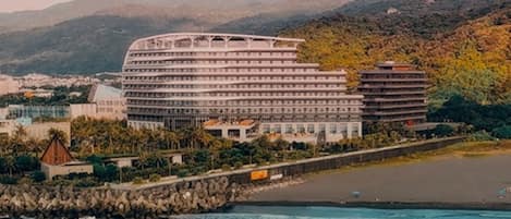 A coastal hotel with multiple floors, a beach in the foreground, and mountains in the background.