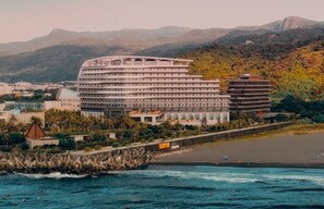 Un hôtel situé sur la côte, avec plusieurs étages, une plage au premier plan et des montagnes en arrière-plan.
