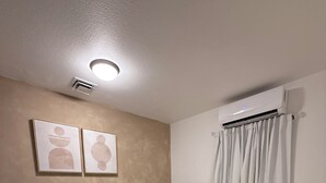 Une chambre à coucher comprenant un lit, une table de chevet, un climatiseur fixé au mur, un luminaire au plafond et deux œuvres d’art murales.