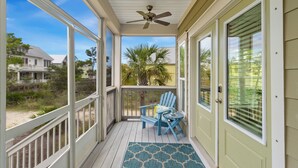 Eine überdachte Veranda mit Deckenventilator, einem blauen Adirondack-Sessel und Blick auf Strand und Häuser im Hintergrund.