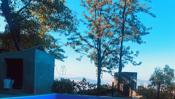 A swimming pool with clear blue water, surrounded by trees and a view of distant hills. - 
Perfect for couples, families, or friends. Book now and unwind in style! (panchgani)