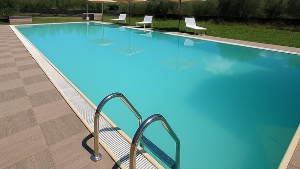 Une piscine rectangulaire avec une terrasse carrelée, un escalier métallique et des chaises longues blanches placées sous des parasols.