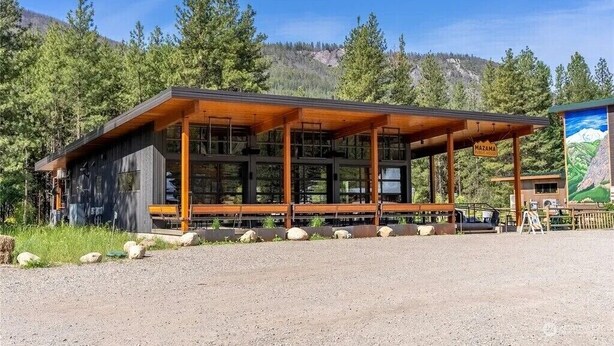 A lodge with a wooden exterior and large windows, surrounded by trees and a mountain backdrop.