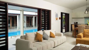 A modern living room with a sofa, a coffee table, and a dining area with wicker chairs. There is a glass door leading to a pool. - Villa 100 - 4BR Private Villa w Private Pool - Fenced Pool (Nadi)
