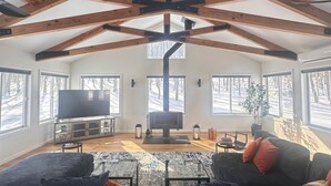 A spacious living room with a high ceiling, exposed wooden beams, a large flat-screen TV, and a fireplace.