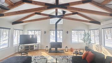 Een ruime woonkamer met een hoge plafond, zichtbare houten balken, een grote flatscreen televisie en een open haard.