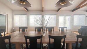 A dining room with a wooden table, six chairs, two pendant lights, and large windows with blinds.