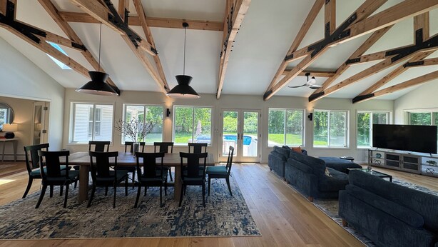 A spacious living room with high ceilings, wooden beams, a dining area with a table and chairs, a large window, and a television.
