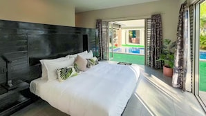 Un grand lit avec une literie blanche et de nombreux oreillers, une tête de lit sombre et une vue sur une piscine, visible à travers des portes coulissantes ouvertes.