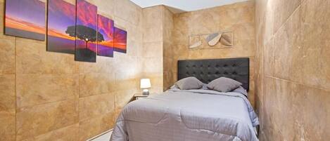 A bedroom with a bed, a nightstand, a wall-mounted lamp, and three framed artworks.