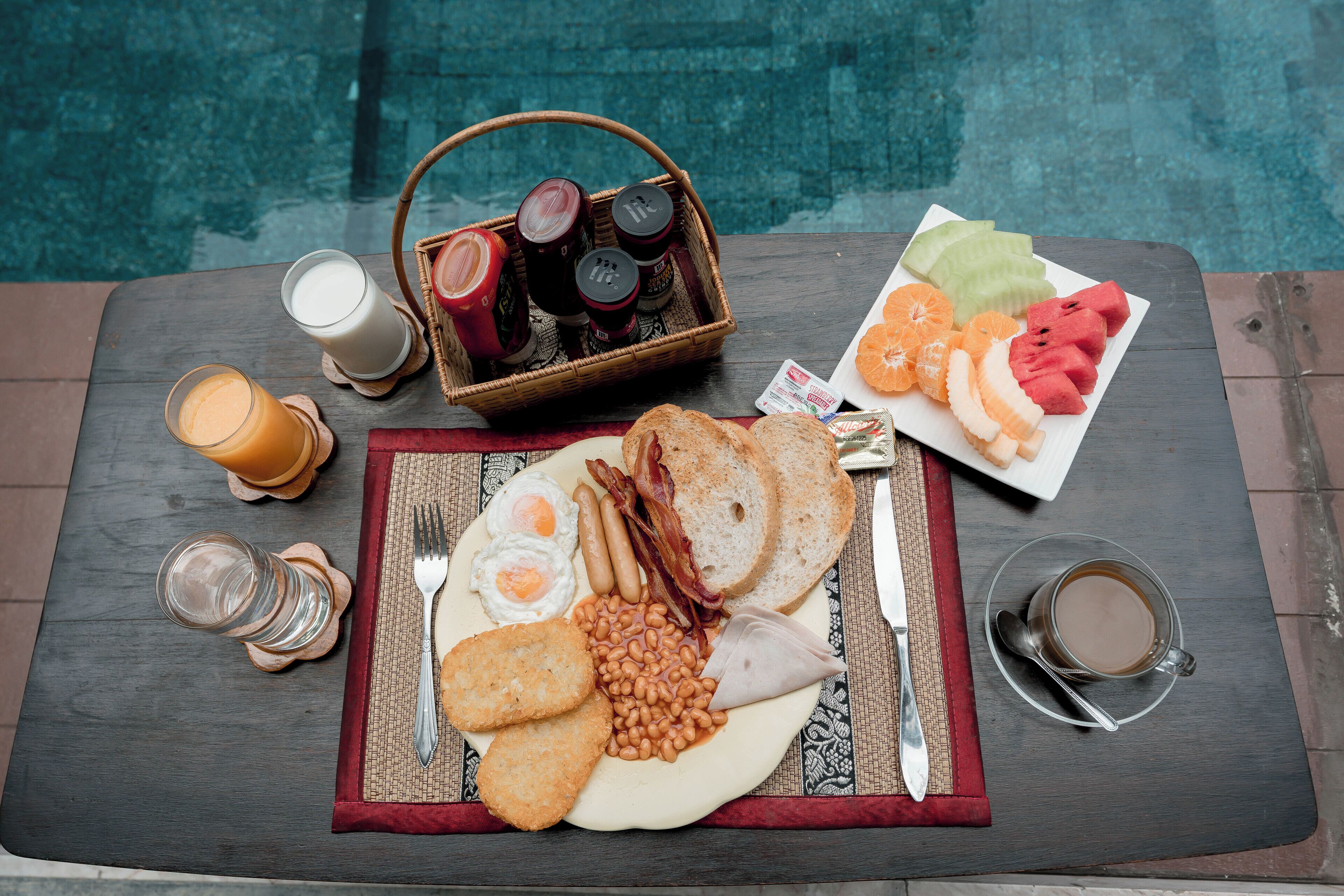 Un desayuno servido en una mesa de madera junto a una piscina, que incluye huevos, tocino, tostadas, frijoles y fruta.