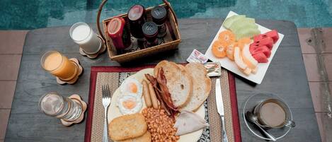 Un desayuno servido en una mesa de madera junto a una piscina, que incluye huevos, tocino, tostadas, frijoles y fruta.