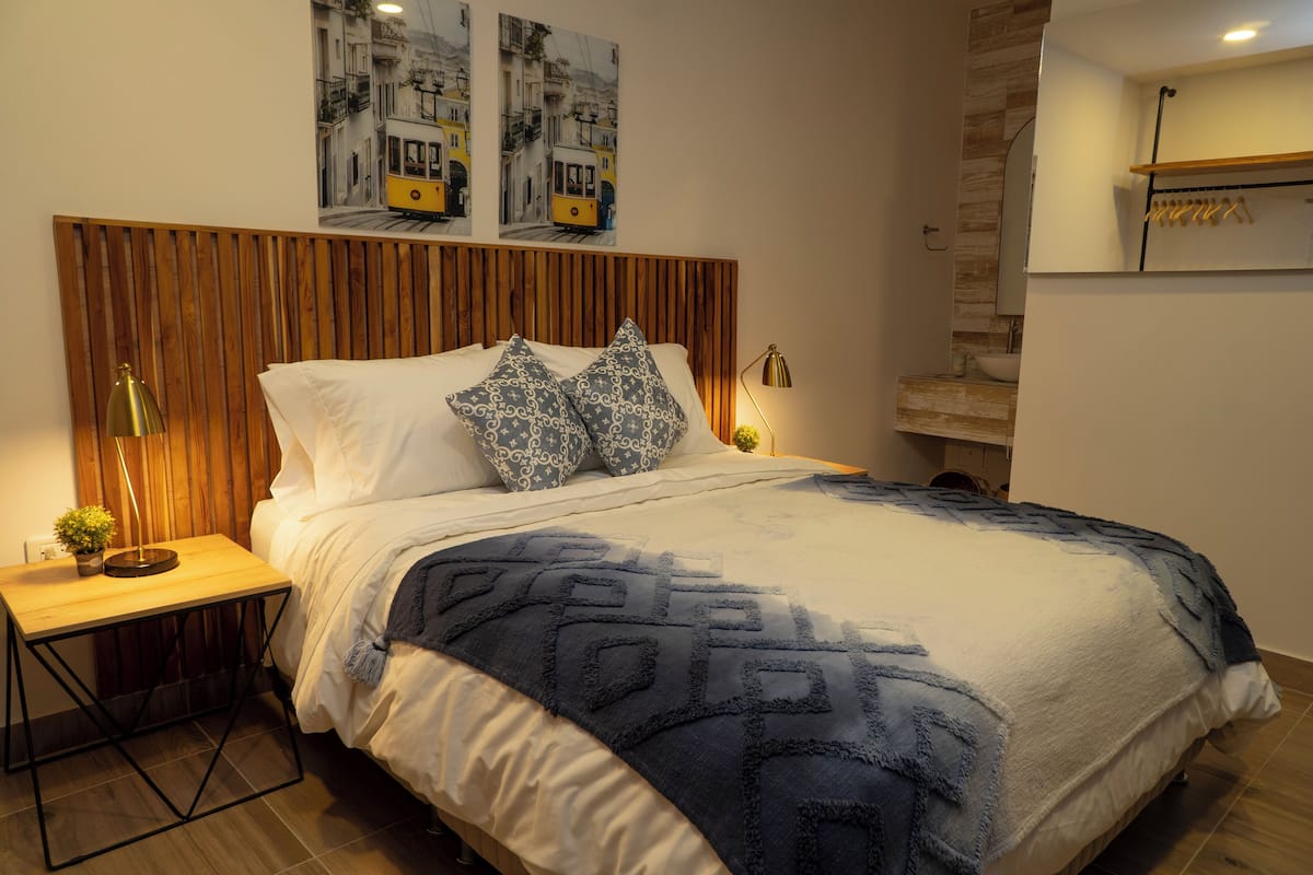 A neatly made bed with patterned pillows, a bedside table with a lamp, and a wall-mounted shelf with clothes.