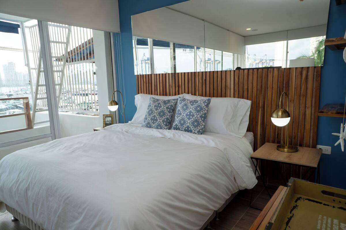 A modern bedroom with a large bed, wooden headboard, and a city view through large windows.