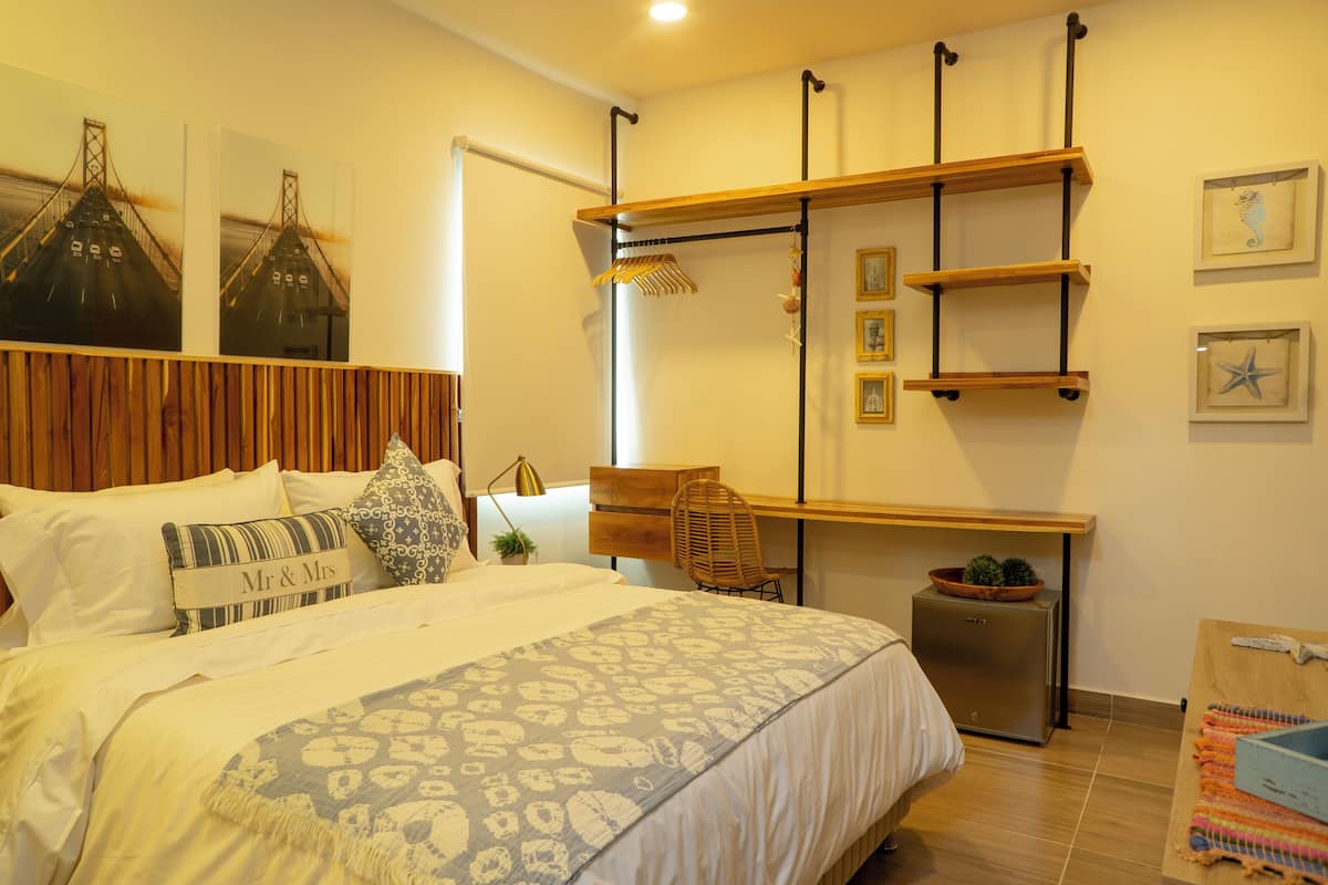 A bedroom with a bed, a desk, a chair, and a small table. There are two paintings on the wall, a potted plant, and a shelf with decorative items.