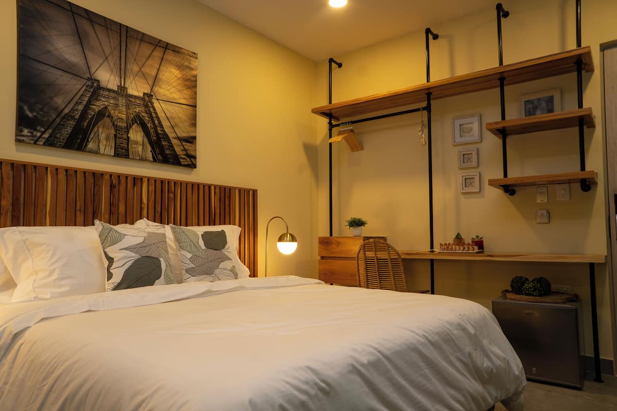 A neatly arranged bedroom with a large bed, a desk with shelves, a lamp, and a wall-mounted picture of a bridge.