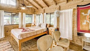 A wooden cabin bedroom with a bed, two chairs, a nightstand, a fan, and a window with curtains. - Edge of the World Cabin & Cabana - 10 minutes to Pacific Ocean & On site (Dominical)