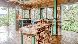 A wooden dining area with a table set for four, a ceiling fan, and large windows offering a view of the surrounding forest. - Edge of the World Cabin & Cabana - 10 minutes to Pacific Ocean & On site (Dominical)