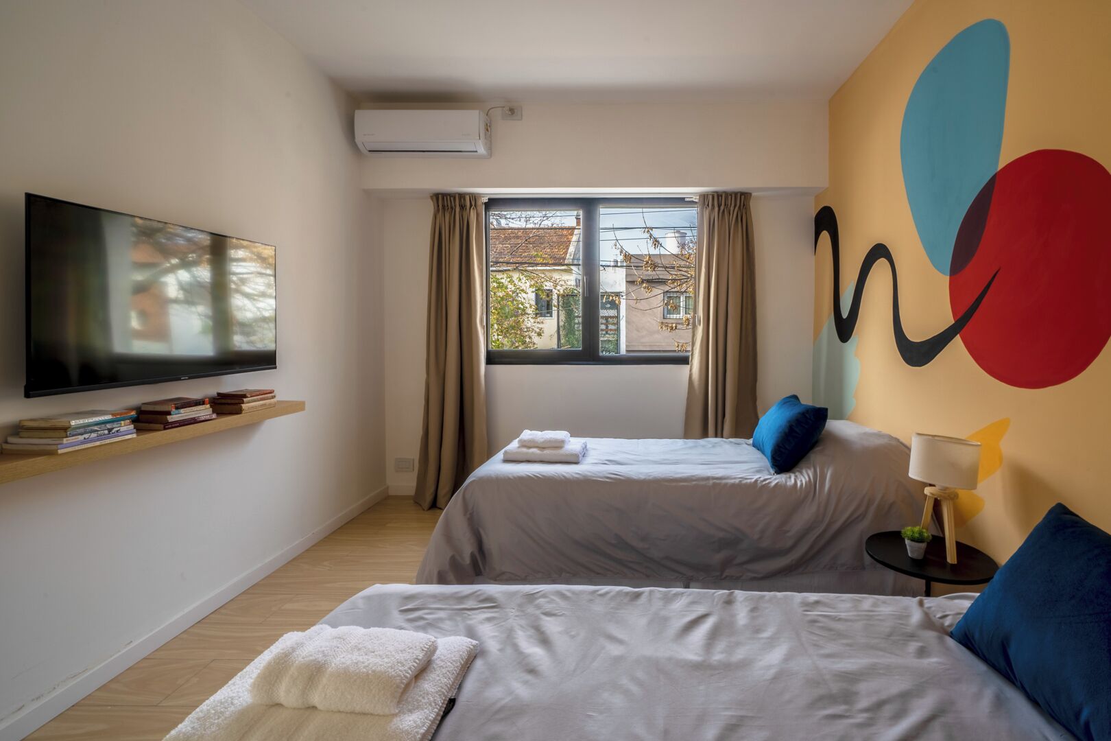 Camera d'albergo con due letti, una TV, una finestra con tende e un'opera d'arte astratta dai colori vivaci.