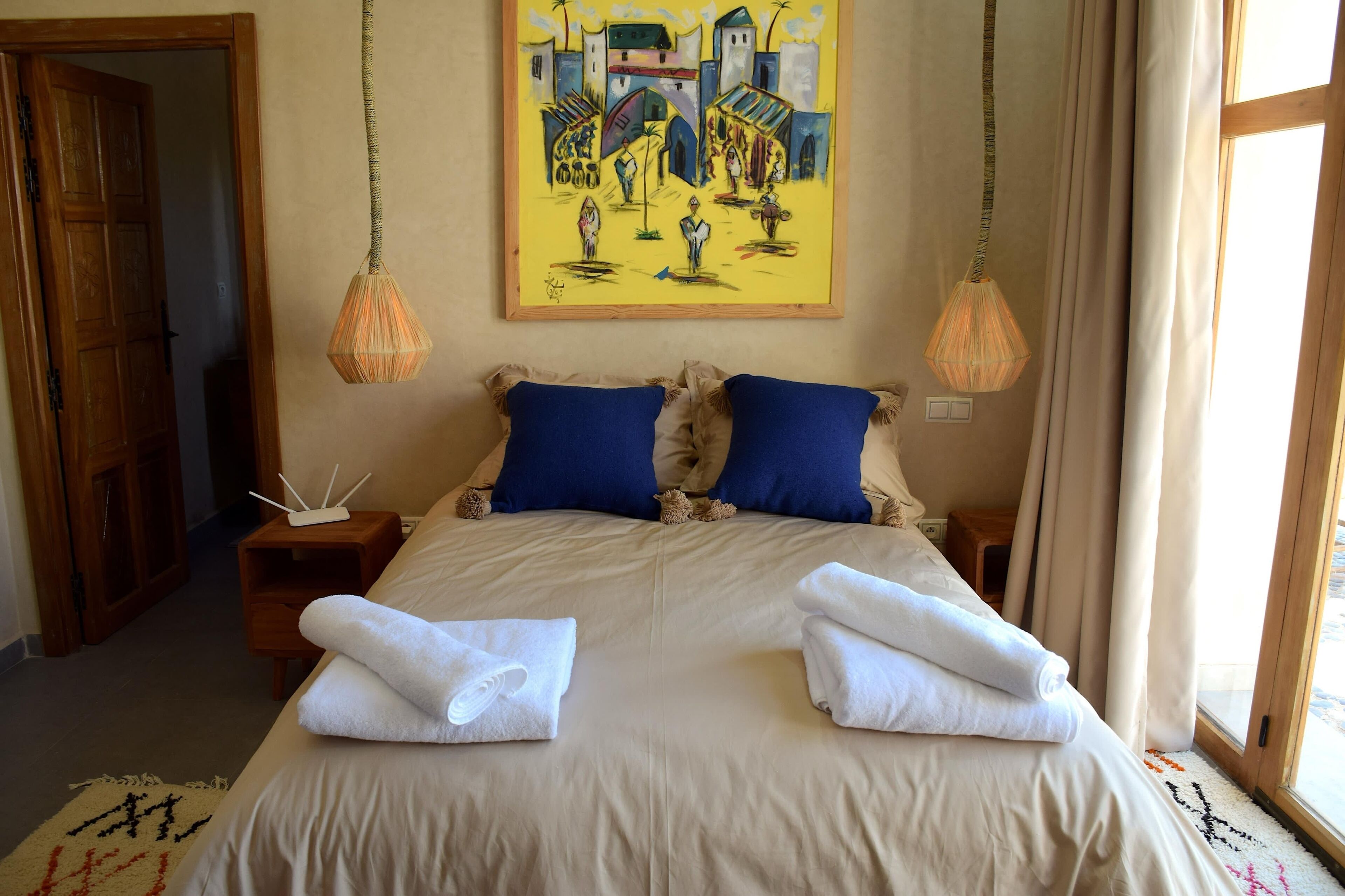 A hotel room with a large bed, two blue pillows, two bedside tables, a painting on the wall, and a window with curtains.
