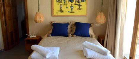 A hotel room with a large bed, two blue pillows, two bedside tables, a painting on the wall, and a window with curtains.