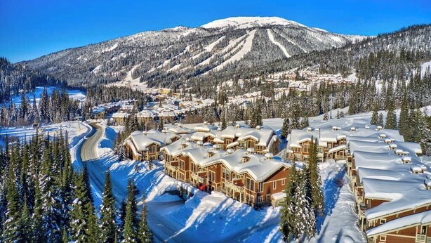 Un complejo turístico en la montaña cubierto de nieve, con edificios de estilo chalet, pistas de esquí y un bosque denso.