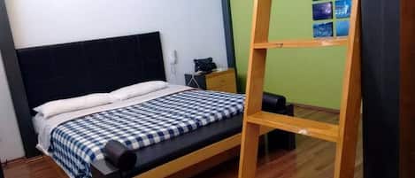 Una cama individual con colcha a cuadros, una mesita de noche de madera con lámpara y un estante en la pared con fotografías.