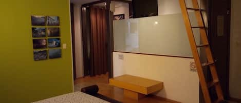 A room with a bed, a small wooden table, a TV mounted on the wall, and a ladder against the wall.