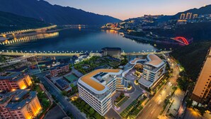 A modern hotel complex with multiple buildings, a central courtyard, and a view of a river and mountains in the background.