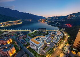 Um complexo moderno de hotéis com vários edifícios, um pátio central e vista para um rio e montanhas ao fundo.